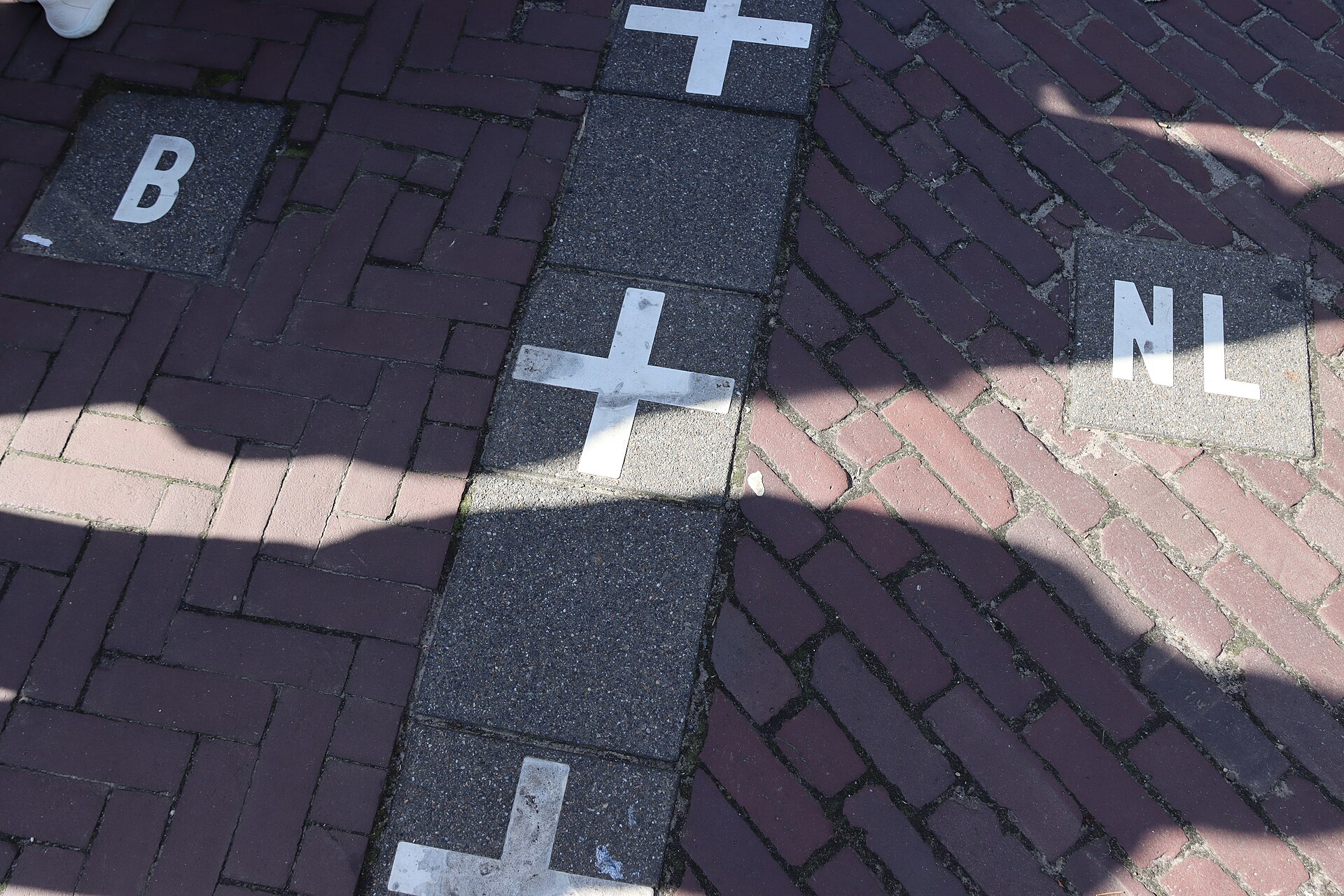 White crosses and 'B' and 'NL' markers set into the brick pavement of a street, showing the Belgium-Netherlands border line at Baarle